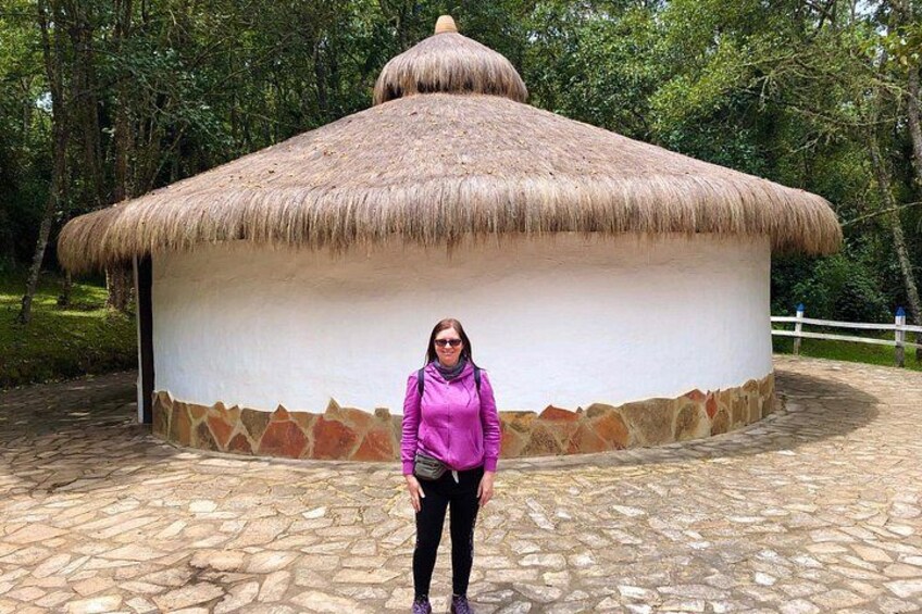A person standing in front of a traditional thatched-roof structure with a stone base, surrounded by a stone-paved area and trees. 5