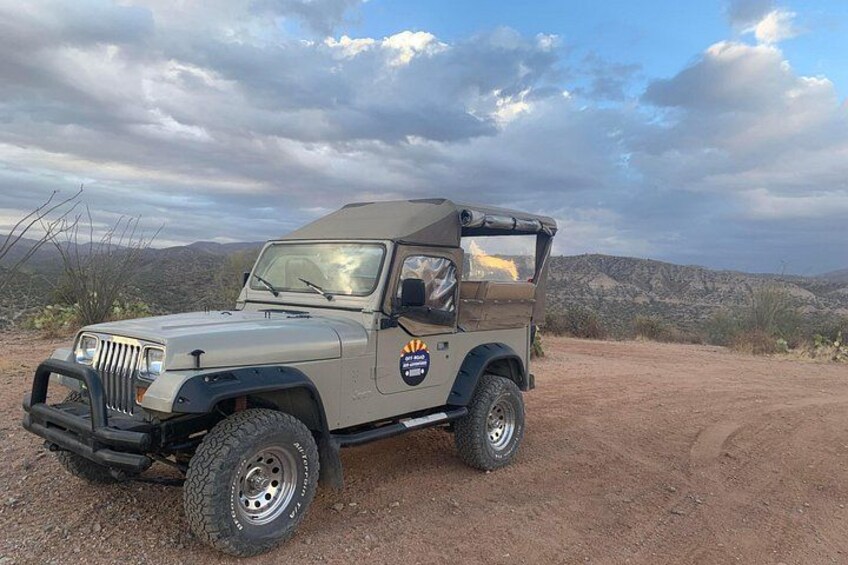 Un vehículo todo terreno color beige con techo, estacionado en un camino de tierra con un paisaje montañoso al fondo. 3
