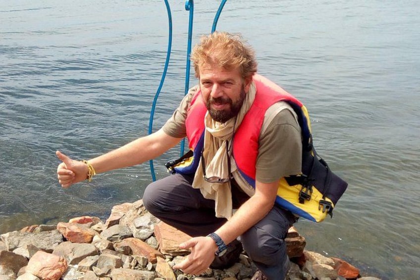 A person sitting on rocks by a body of water, wearing a life jacket and a backpack. 1