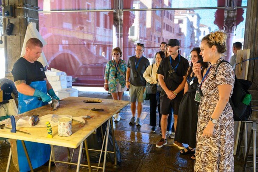 A person in a patterned dress speaking to a group of people in an industrial setting with a workbench and tools. 4