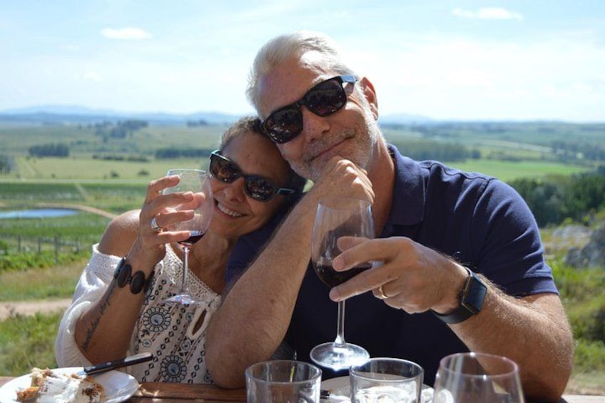 Deux personnes trinquent avec des verres de vin à une table, avec en arrière-plan une vue pittoresque sur la campagne. 2