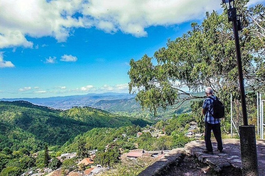 A man standing on a rocky outcrop overlooking a valley with dense greenery and distant hills. 2