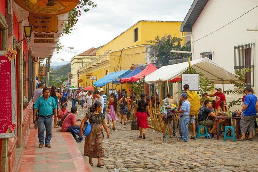 A cobblestone street with people walking, sitting at outdoor tables, and colorful umbrellas. 3