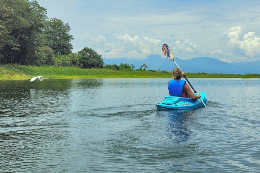 A person kayaking on a calm body of water with greenery and mountains in the background. 2