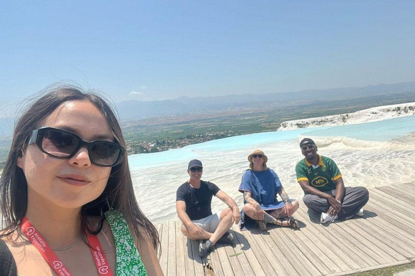 A group of people sitting on a wooden deck overlooking a large, clear blue pool with a mountainous landscape in the background. 4
