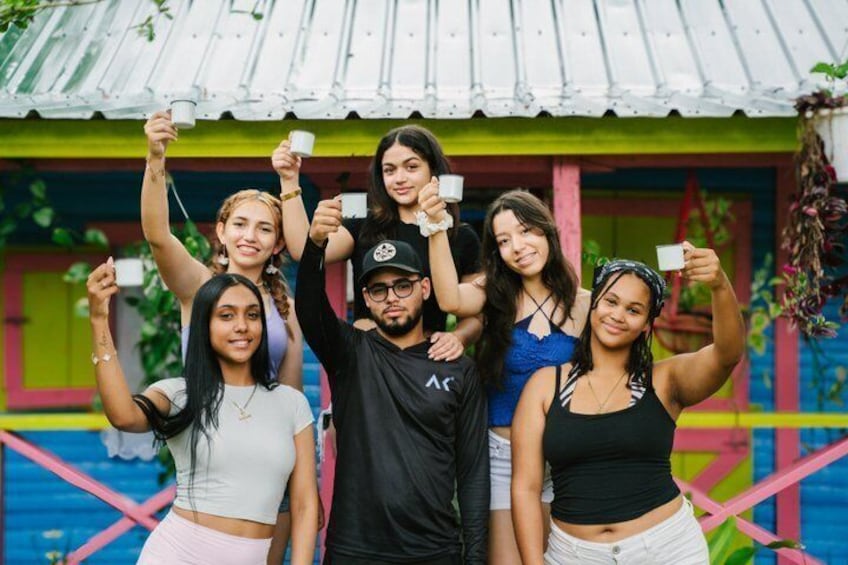 A group of people holding up white cups in front of a colorful house with a corrugated metal roof. 4