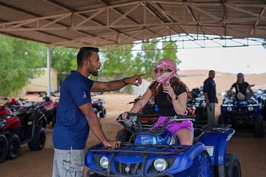 Una persona señalando un ATV azul, otra persona sentada en él, y un área techada con ATVs estacionados. 2