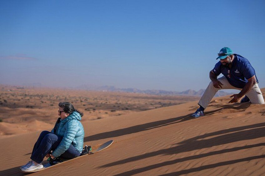 Zwei Personen sitzen auf einer Sanddüne mit einem Skateboard, eine Wüstenlandschaft und ein klarer Himmel im Hintergrund. 3