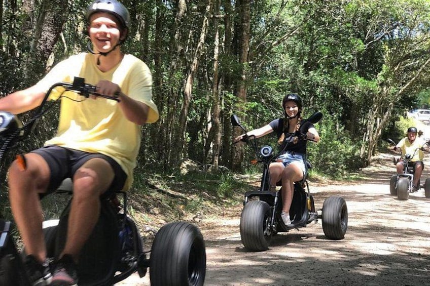 People riding three-wheeled vehicles on a dirt path surrounded by trees. 1