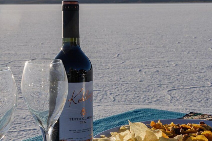 Una botella de vino Tinto Classico 2022, dos copas de vino vacías y un plato con comida sobre una superficie nevada. 3