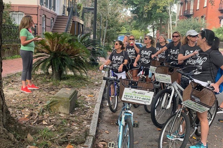 A group of people on bicycles, a woman speaking to them, and a sign for a bike rental service. 1