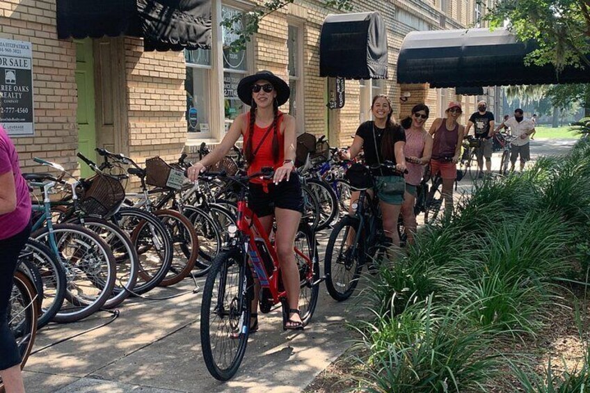 A group of people riding bicycles on a sidewalk, with a building and parked bicycles in the background. 5