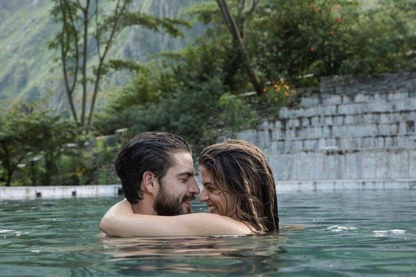 Dos personas abrazándose en el agua, con un muro de piedra y follaje al fondo. 3