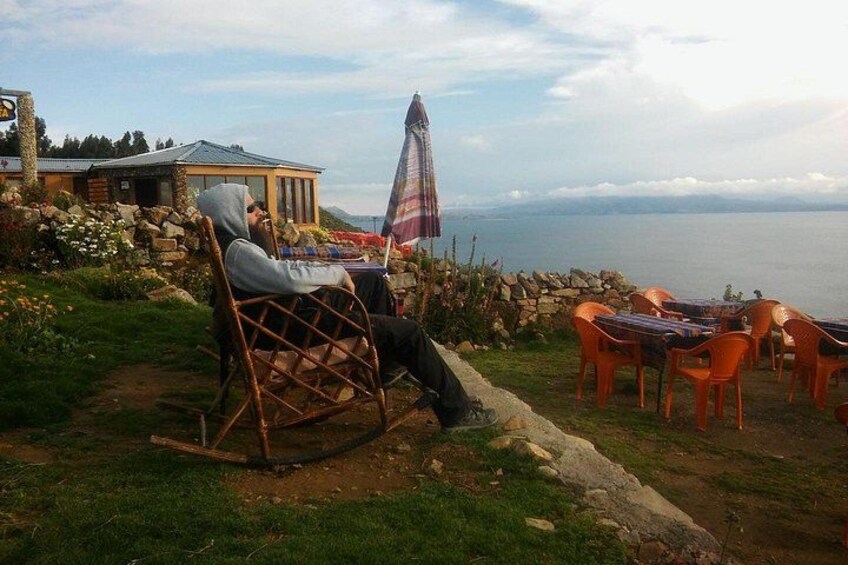 A person sitting on a rocking chair overlooking a scenic view with a table and chairs, a stone wall, and a building in the background. 2