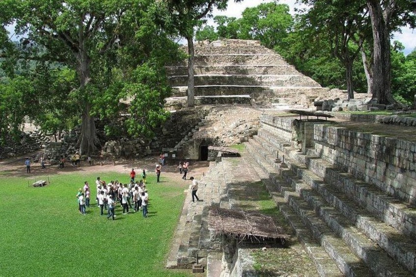 A large stone pyramid with a staircase leading up to it, surrounded by trees and a grassy area with people gathered around. 2