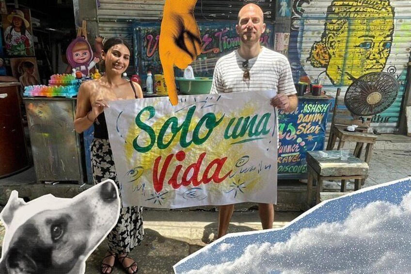 A person holding a sign with the text 'Solo una Vida' in a street setting with graffiti and a fan in the background. 4