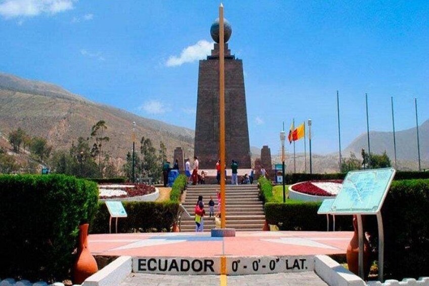 A monument with a flagpole, a sign indicating Ecuador's latitude, and a mountainous landscape in the background. 2