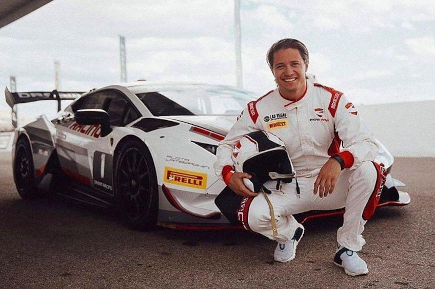 A white racing car with sponsor logos, a person in racing gear, and a bridge in the background. 5