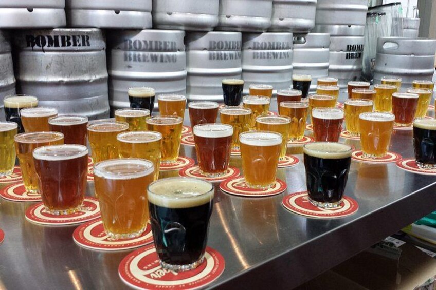 Un bar con varios vasos de cerveza, algunos con espuma, y varias pilas de barriles con la etiqueta 'Bomber Brewing' al fondo. 3