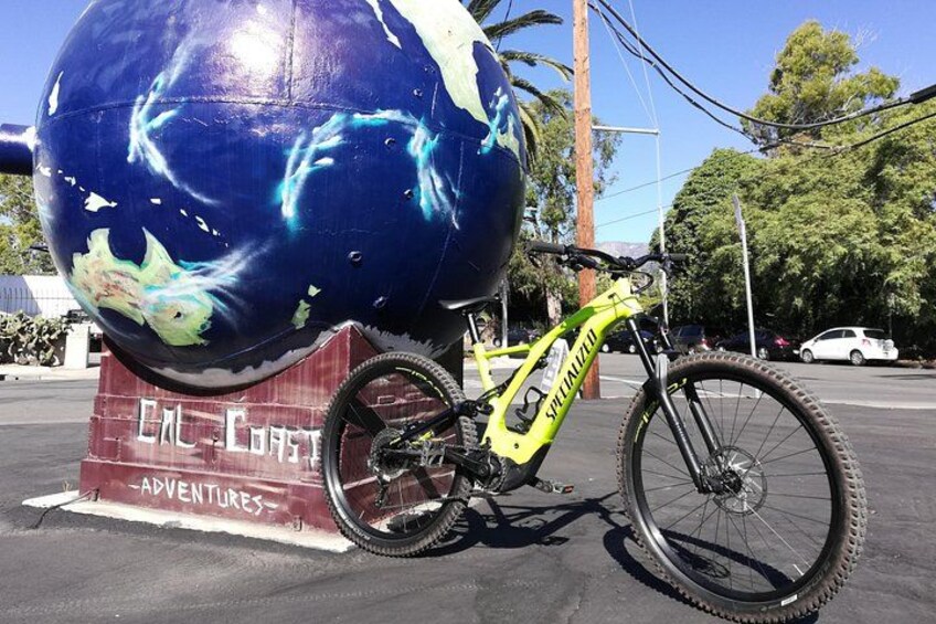 Een gele mountainbike geparkeerd naast een groot sculptuur van een wereldbol met een blauw-wit ontwerp, op een parkeerplaats met auto's en bomen op de achtergrond. 3