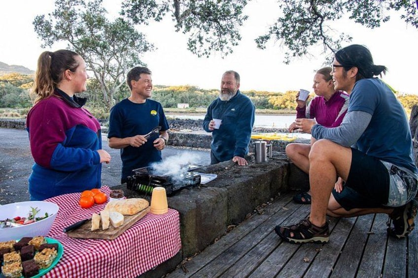 A group of people around a barbecue grill outdoors with a checkered tablecloth, food, and a scenic background. 3