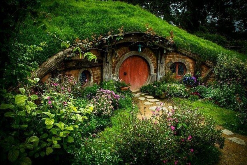 A Hobbiton-style house with a red door, surrounded by lush greenery and flowering plants. 2
