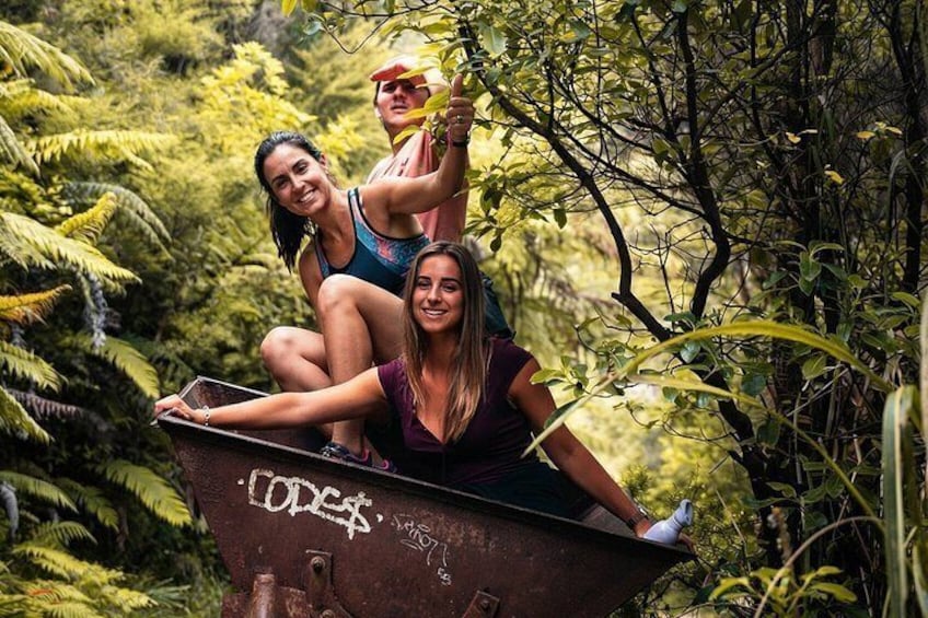 Three people sitting on a rusty barrel in a forested area with dense greenery. 1