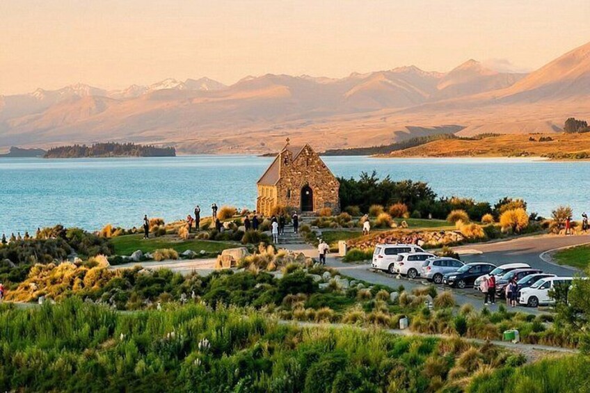 A stone church by a lake with mountains in the background and parked cars in the foreground. 3
