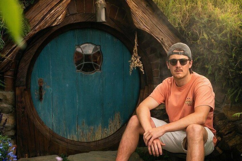A person sitting in front of a blue door with a circular window, surrounded by greenery. 4
