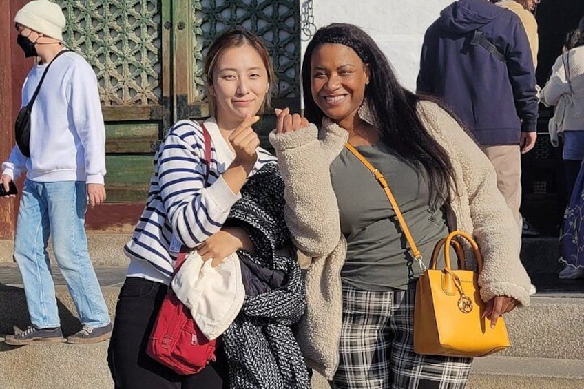 Two people posing for a photo, one wearing a striped shirt and the other in a beige jacket, with a green gate and a yellow bag in the background. 3