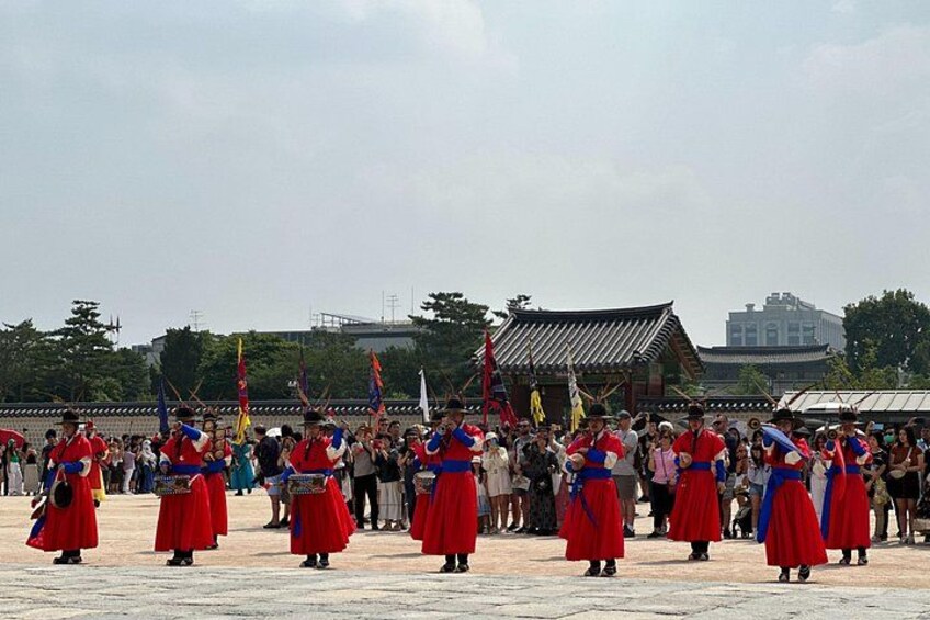 A group of people in traditional red and blue attire performing a dance in an open area with a traditional building in the background. 1