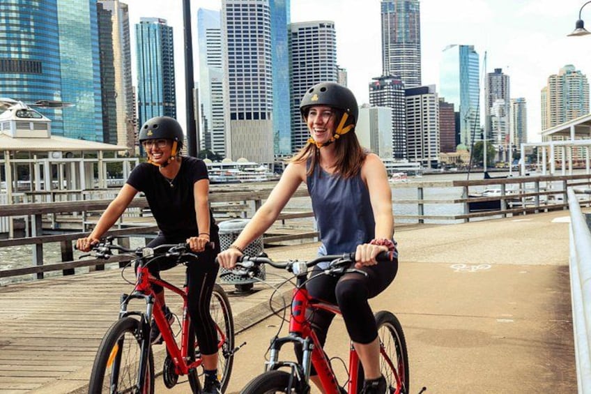 Two people riding bicycles on a bridge with a city skyline in the background. 3