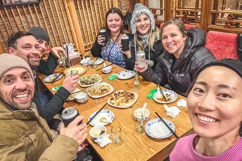A group of people sitting around a wooden table with plates of food, holding drinks, and smiling. 1