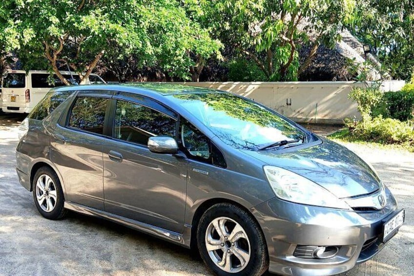 A silver car parked on a concrete surface with trees and a white van in the background. 1