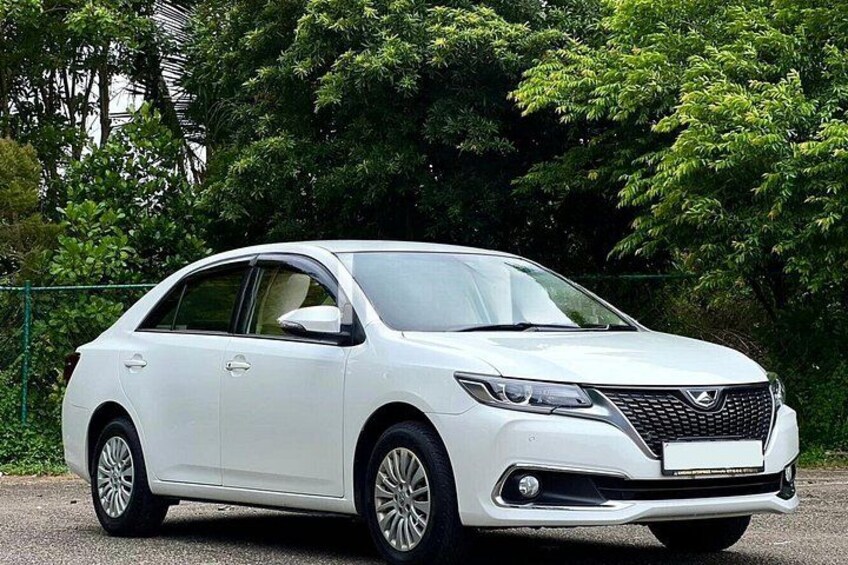A white sedan parked on a paved surface with greenery in the background. 1