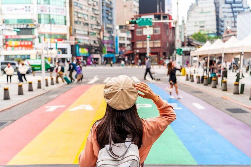 A person walking on a rainbow-colored path in an urban setting with buildings and pedestrians in the background. 5