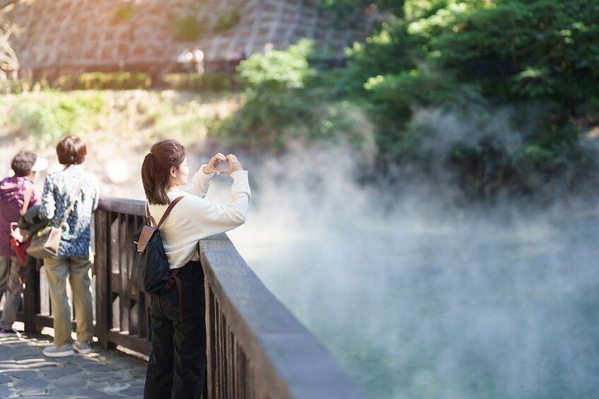 A person taking a photo of a steam vent, with a bridge and greenery in the background. 2