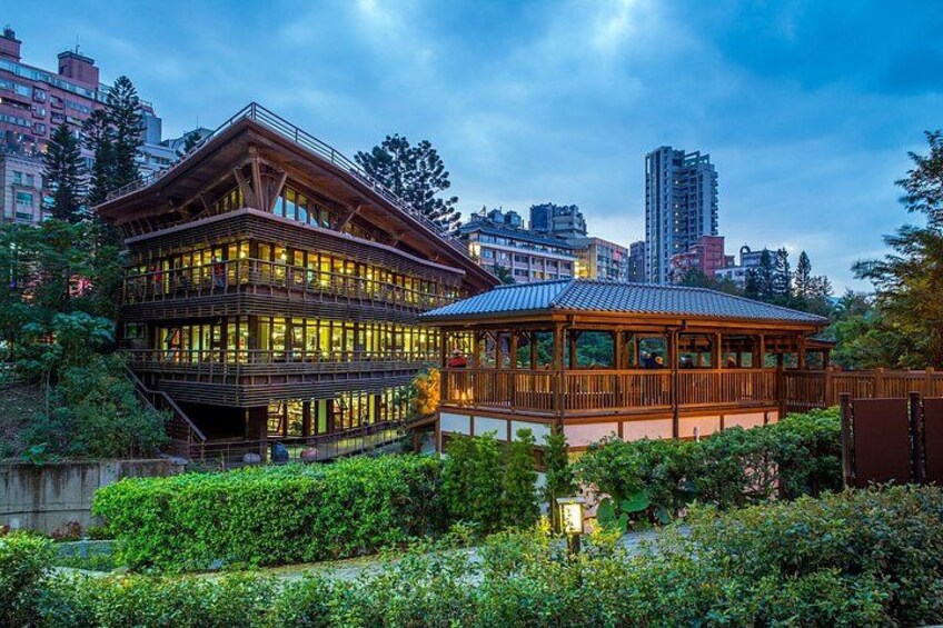 A traditional wooden building with a glass facade, surrounded by lush greenery and a cityscape in the background. 3