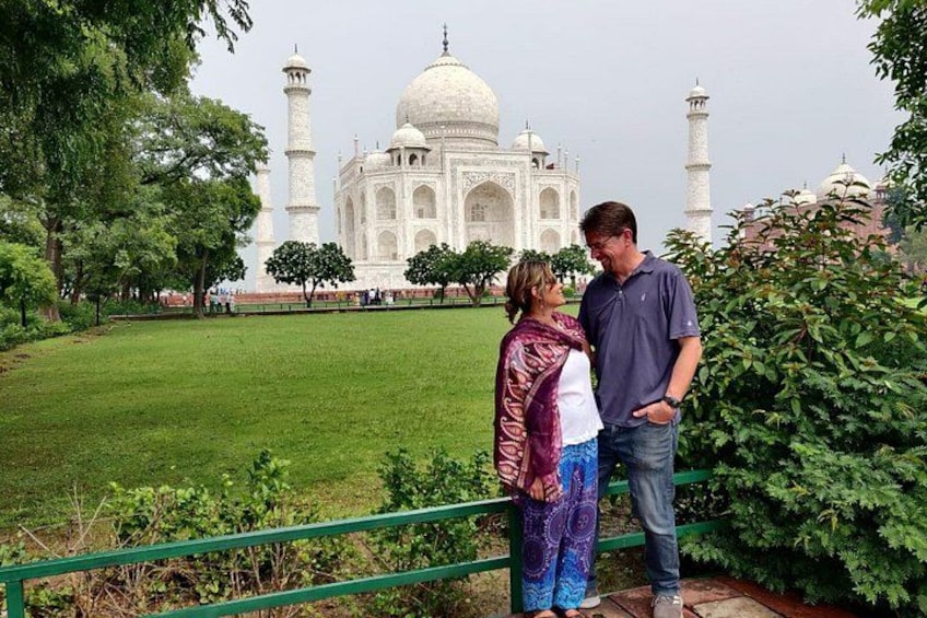 Una pareja frente al Taj Mahal, con una cerca verde y árboles en primer plano. 4