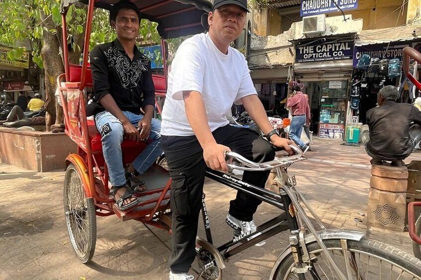 Un hombre montando en bicicleta con un pasajero en un rickshaw, una tienda con un letrero que dice 'THE CAPITAL OPTICAL CO.', y una estatua al fondo. 3