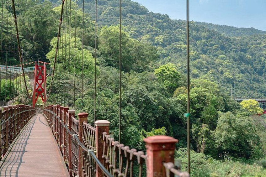 A suspension bridge with red railings and a wooden walkway, surrounded by dense green forest and hills. 1
