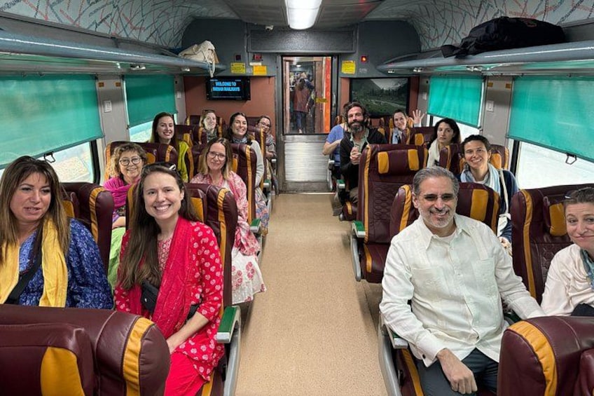 Interior de un tren con pasajeros sentados, una pantalla de televisión y una ventana que muestra un paisaje. 1
