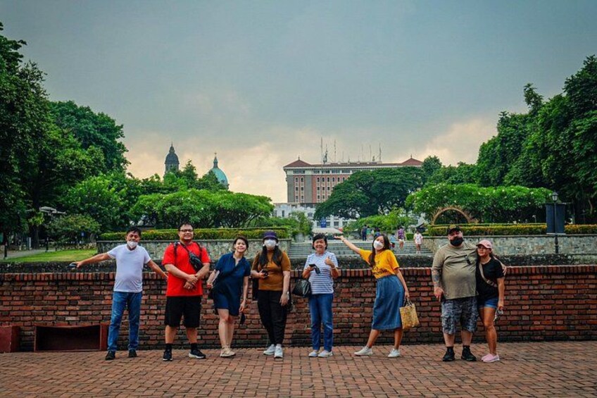 A group of people standing in a park with a brick wall and trees in the background. 3
