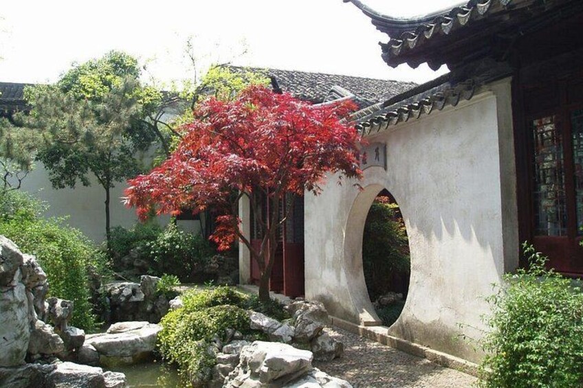 Un patio tradicional con un árbol de hojas rojas, un puente de piedra y un edificio blanco con techo de tejas. 2