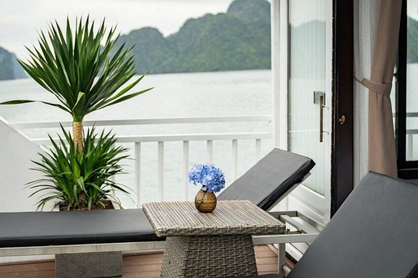 A balcony with lounge chairs, a small table with a vase of flowers, and a view of a body of water with mountains in the background. 2