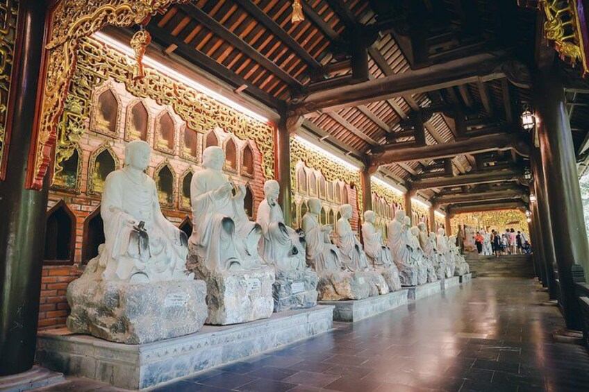 A series of stone statues in a covered corridor with intricate wooden architecture. 4