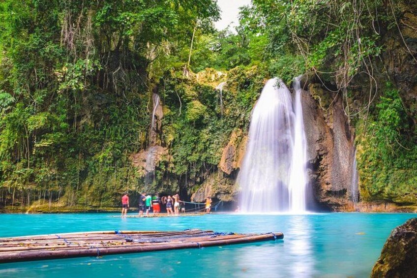 Ein Wasserfall, ein klares, blaues Becken und eine Gruppe von Menschen auf einer schwimmenden Plattform. 5
