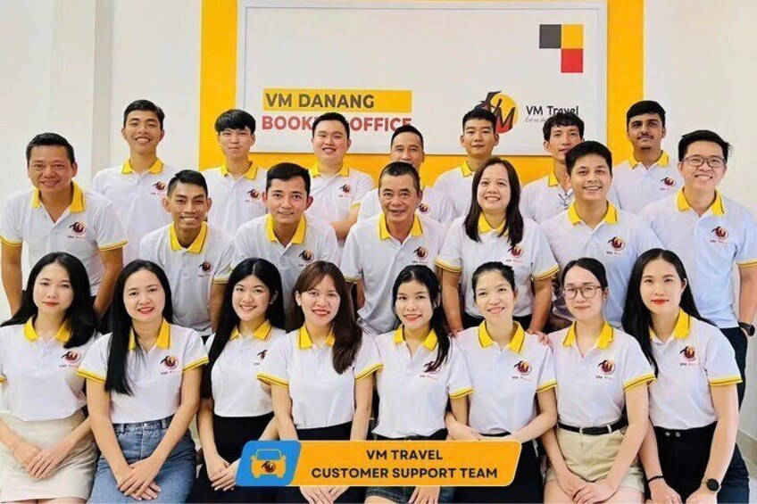 A group of people in a meeting room with a banner reading 'VM DANANG BOOK OFFICE' and 'VM Travel Customer Support Team'. 2
