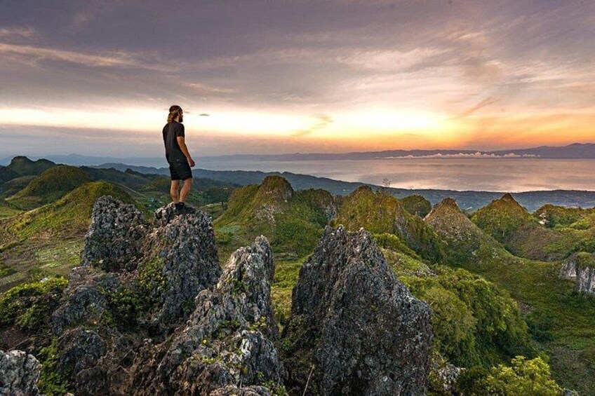 A person standing on a rocky outcrop overlooking a mountainous landscape with a view of the ocean at sunset. 5