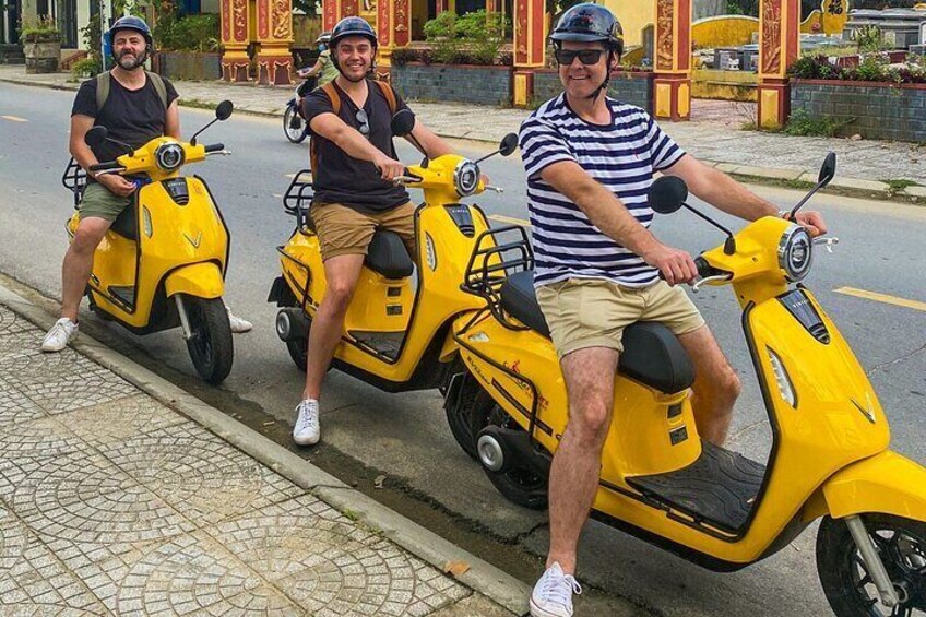 Three people riding yellow scooters on a paved road with decorative buildings in the background. 5